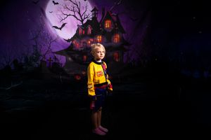 Child in yellow costume stands against dark purple Halloween backdrop with haunted house and full moon.