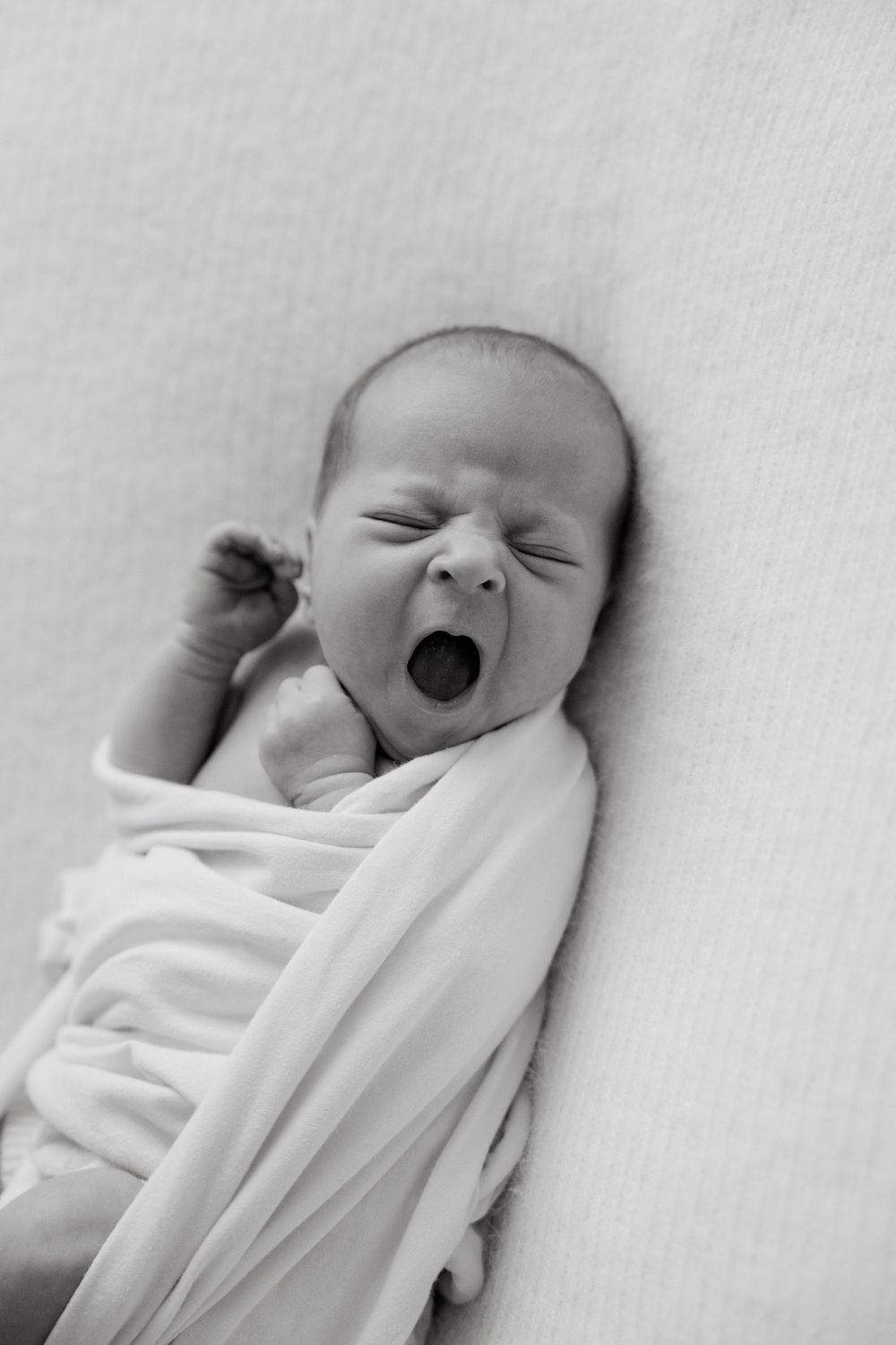 Black and white photo of a newborn baby swaddled in white fabric expressing joy with a big smile.