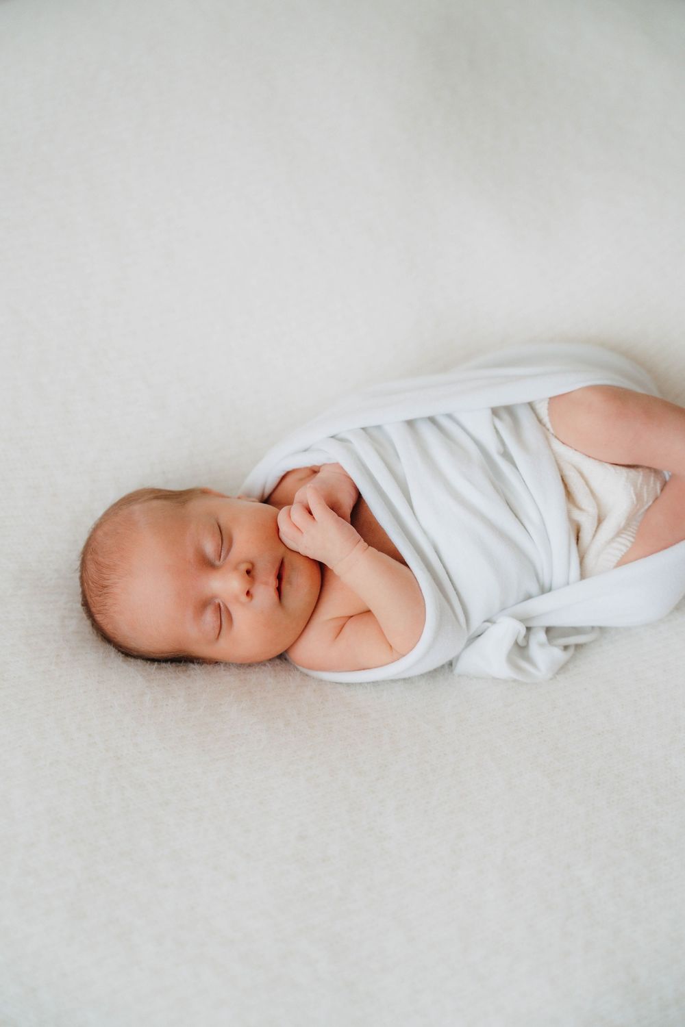 Sleeping infant wearing white onesie lying on light colored surface in gentle natural light.