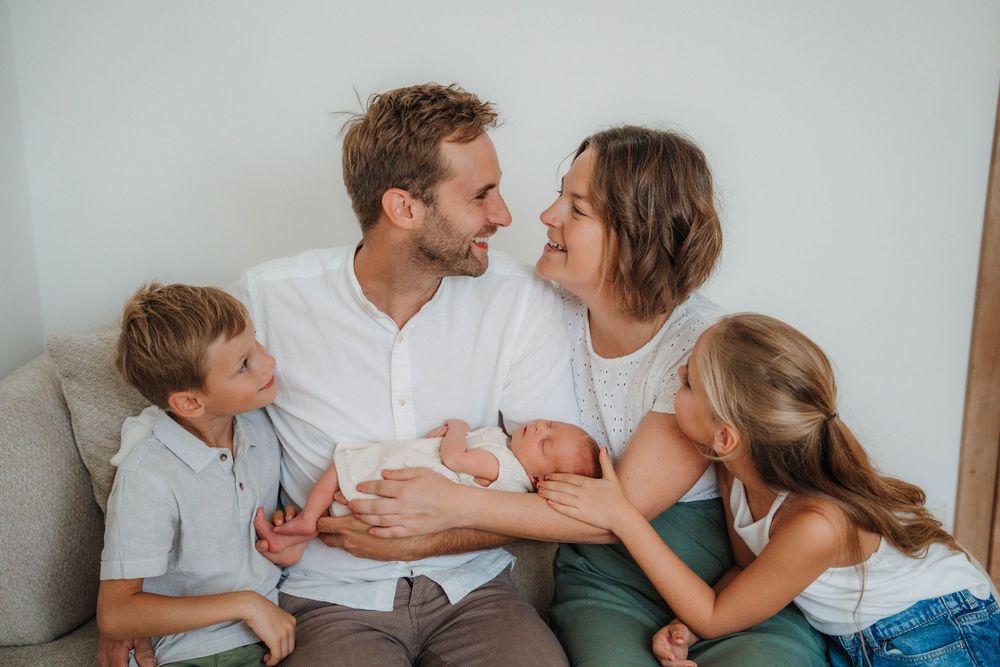 A family of four sits together on a couch sharing an intimate moment with their newborn baby.
