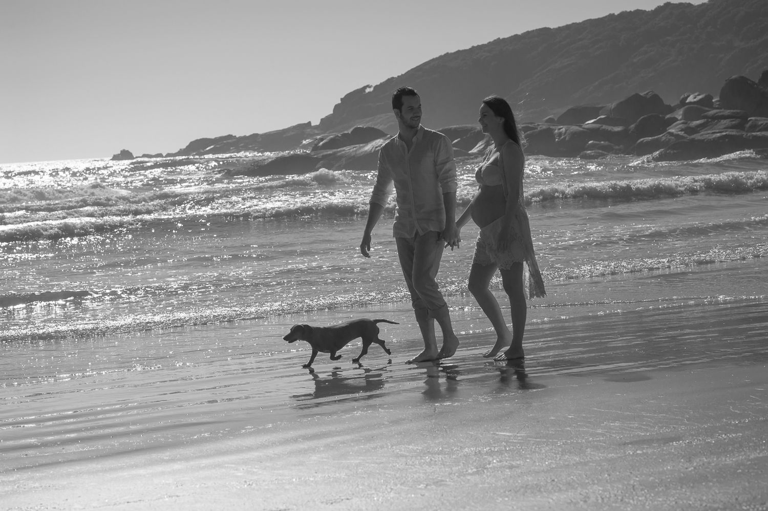 A couple walks along the beach with their small dog as waves crash behind them.