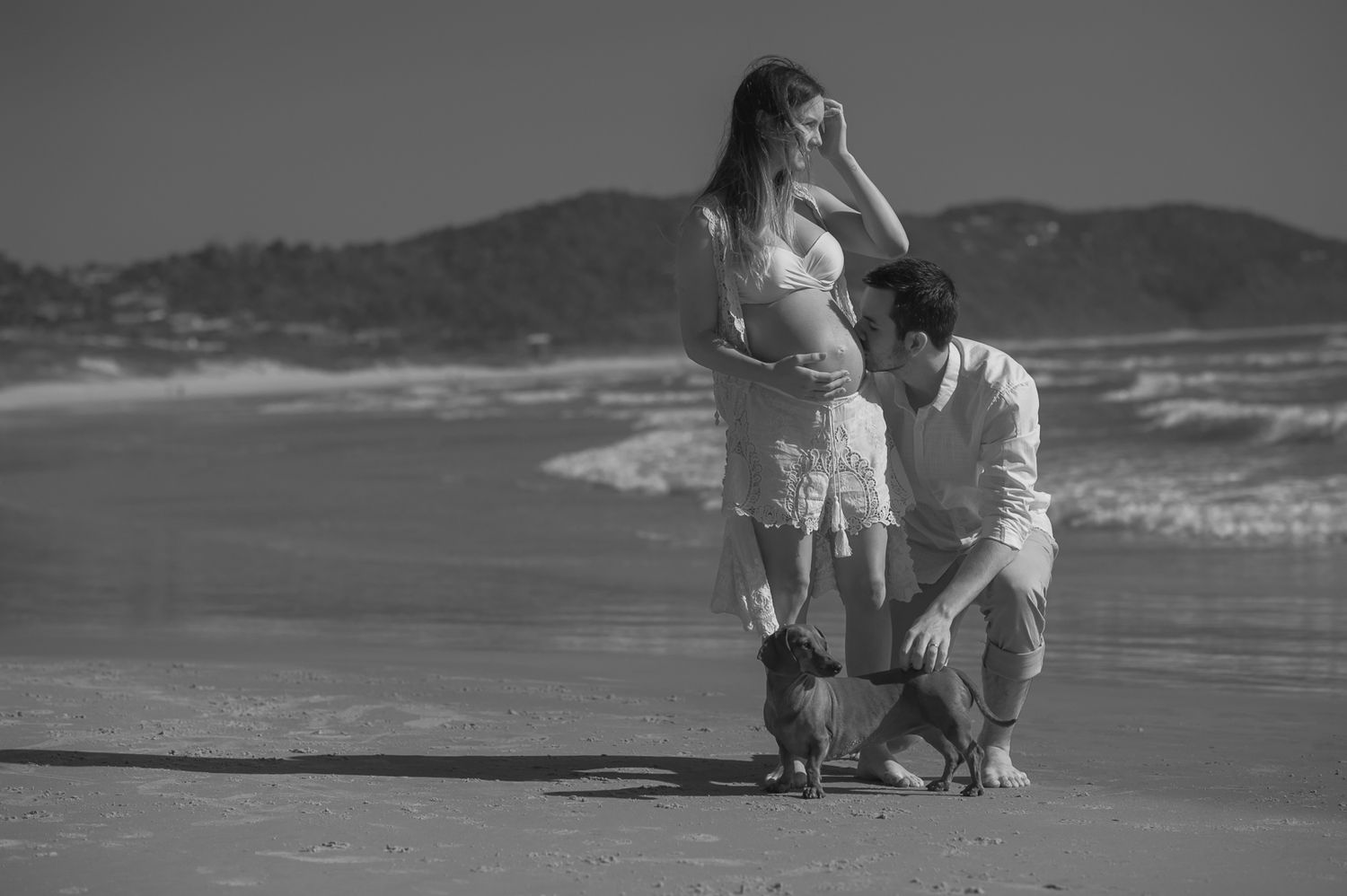 A tender moment shared between two people on a beach with waves and mountains in the background.