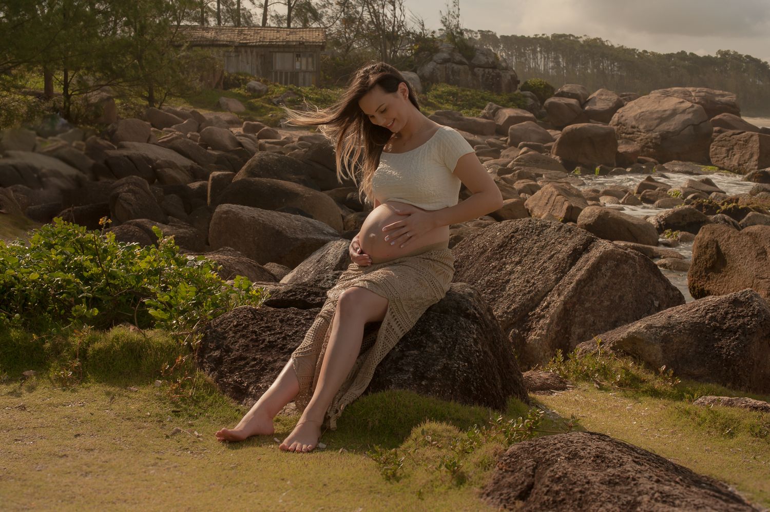 Person in casual attire sitting on rocky coastal terrain during golden hour.