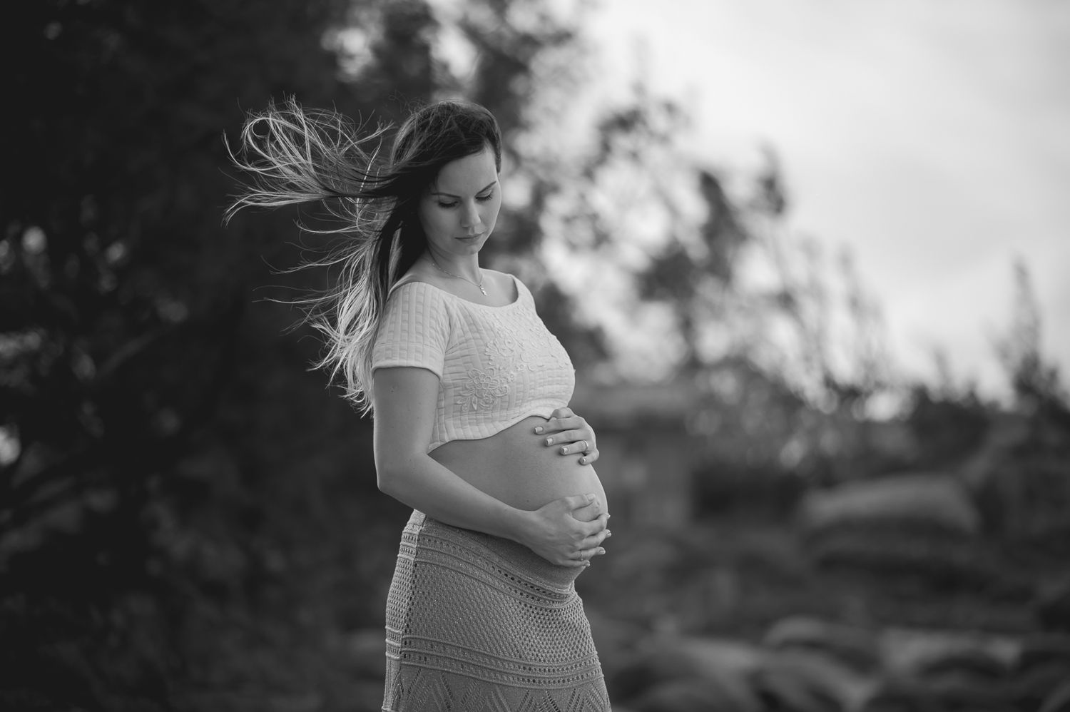 An expectant mother in a white top poses against a nature backdrop with windswept hair.