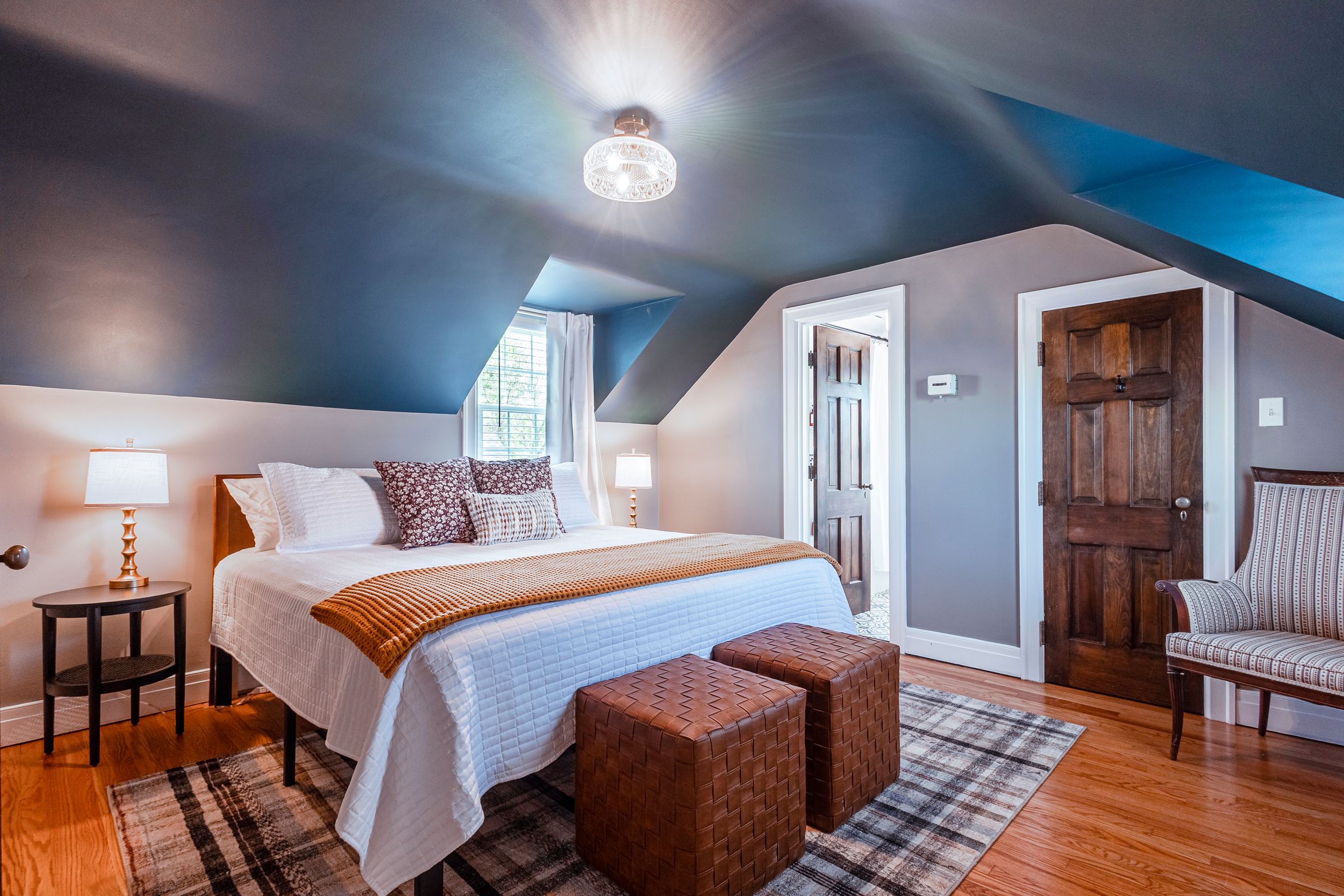 Cozy attic bedroom with blue walls, white bedding, brown accents and plaid area rug under sloped ceiling.