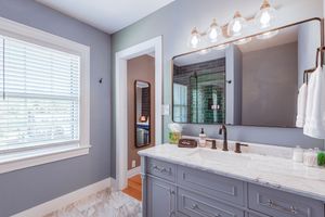 Gray bathroom with marble countertop, framed mirror, and white vanity cabinet beneath window.