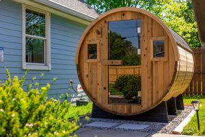 Wooden barrel sauna installed beside a light blue house with landscaping.