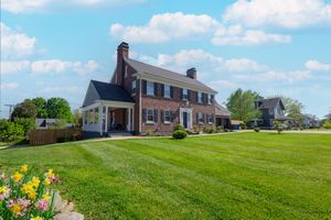 Stately brick colonial house with side porch and well-maintained landscaping under blue sky.
