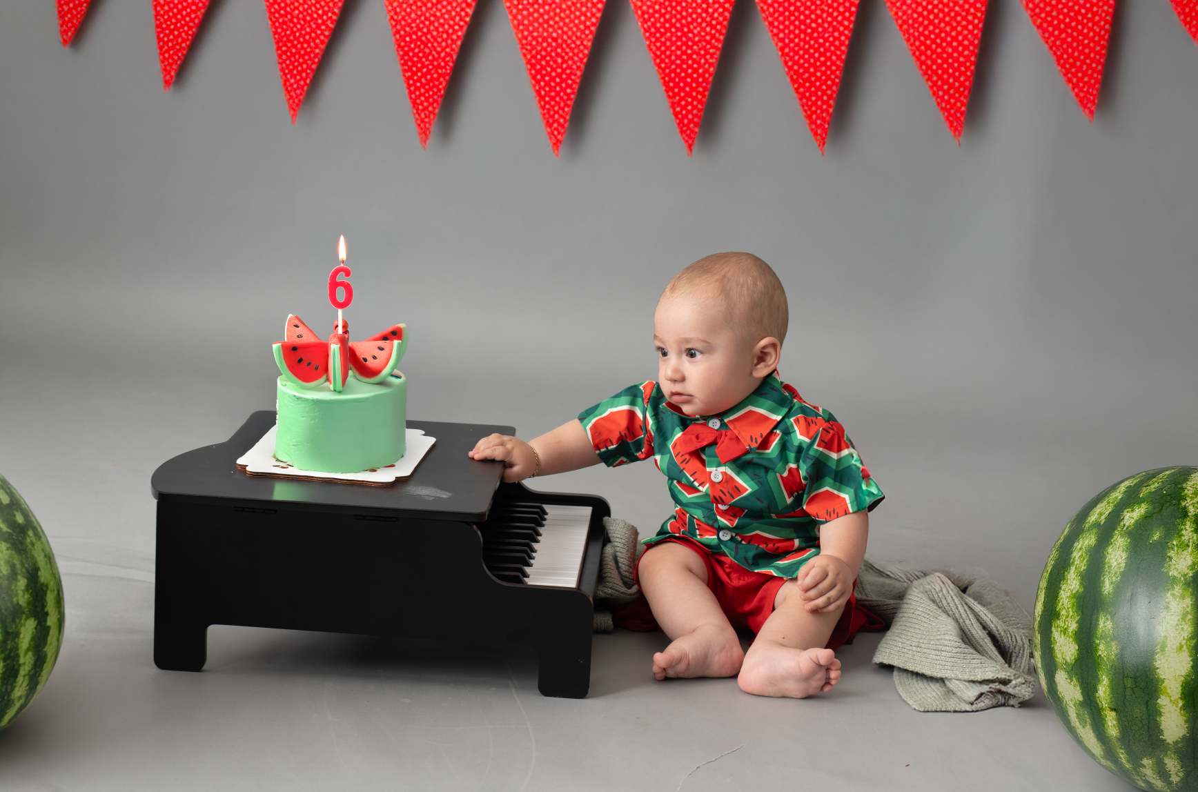 Baby in watermelon outfit touches a tiny piano beside a green birthday cake, surrounded by watermelons on a gray backdrop.