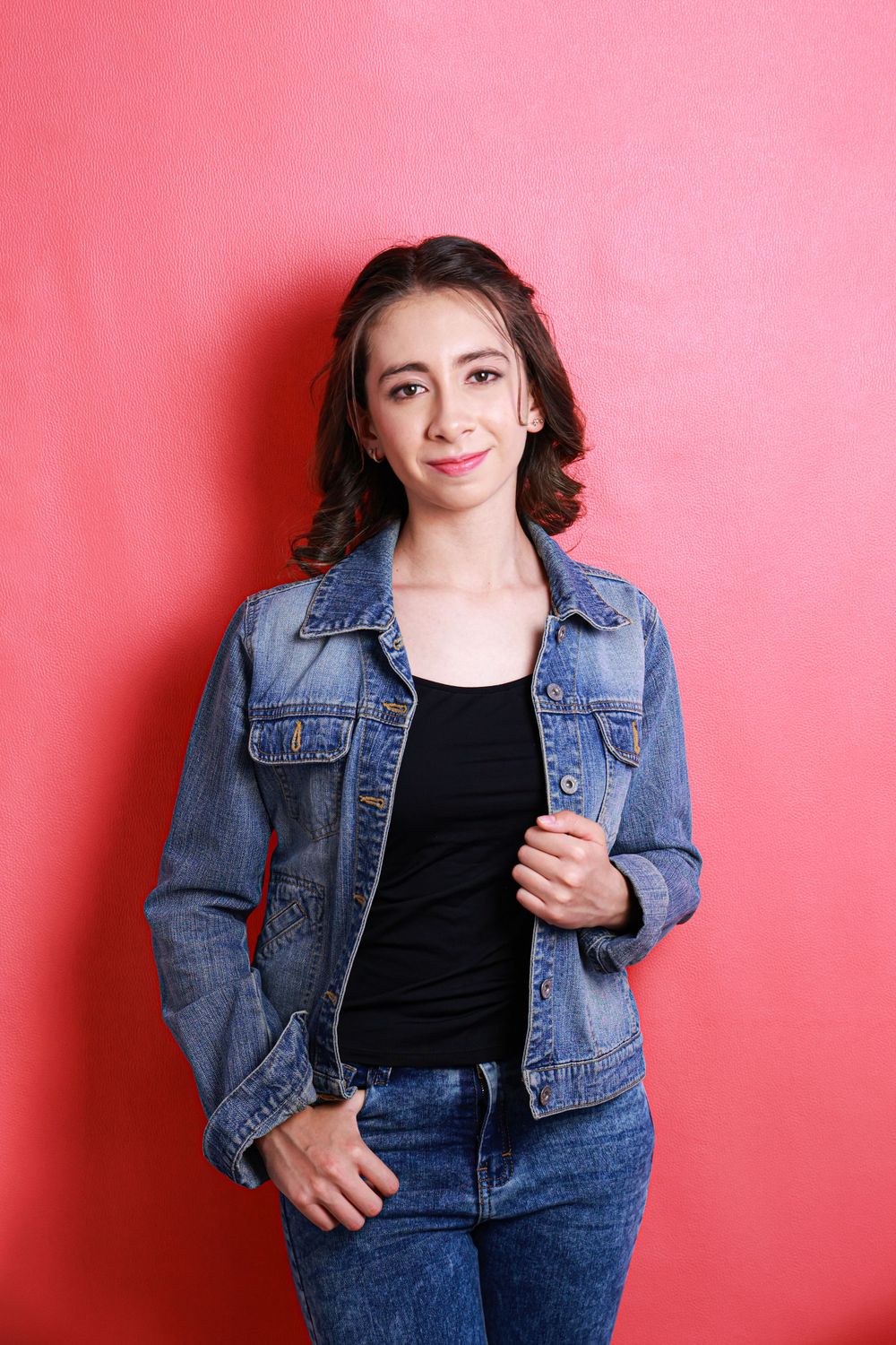 Fashion model in denim jacket and black top poses against pink backdrop in casual style photoshoot.