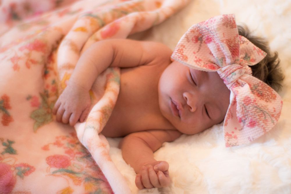 Adorable newborn baby girl sleeping peacefully wearing a floral pink bow headband on a soft blanket.