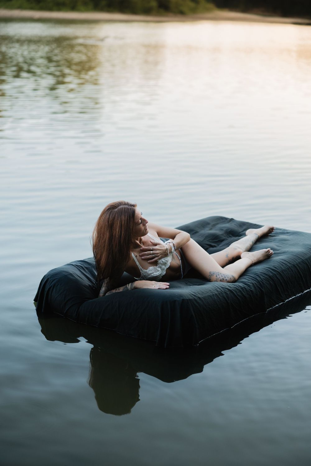 Woman relaxing on a black inflatable mattress floating on a calm lake at sunset.