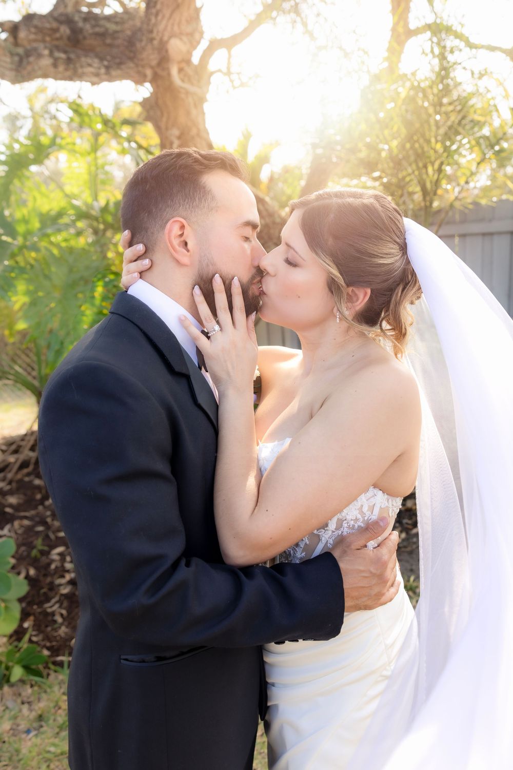 Bride and groom share a romantic kiss outdoors, with soft golden sunlight filtering through trees in the background.