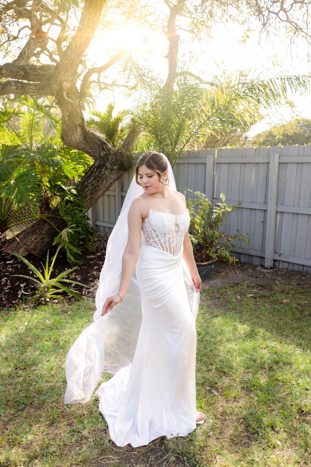 Bride in white strapless lace wedding gown with veil poses in sunlit garden near tropical plants and wooden fence.