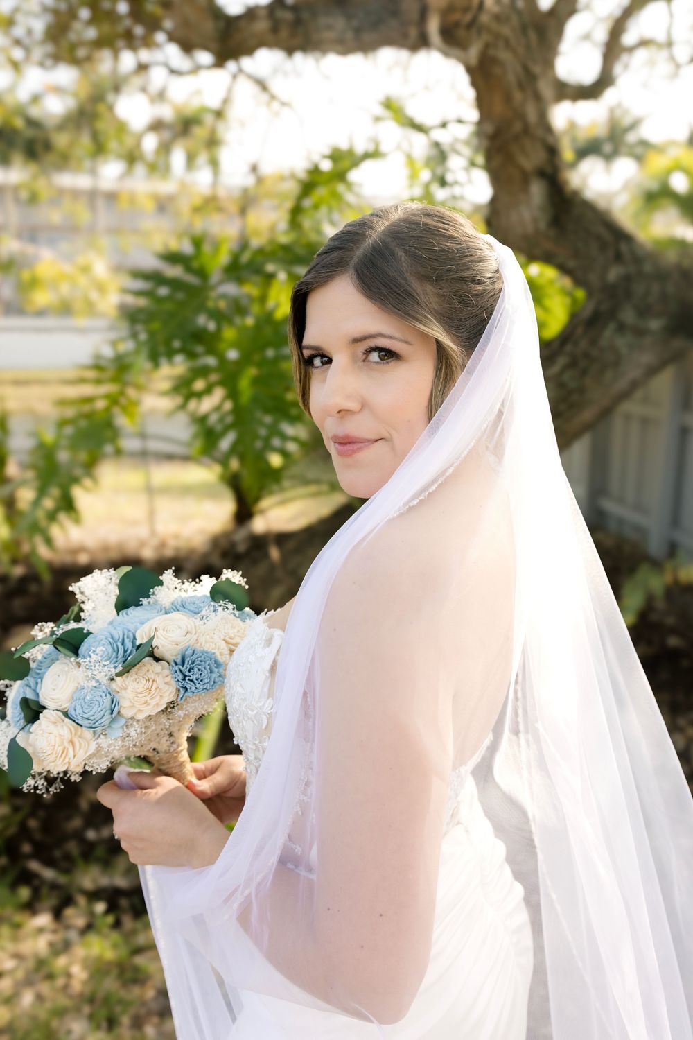 Bride in white gown and veil holds blue and white bouquet, posing outdoors near a tree on her wedding day.