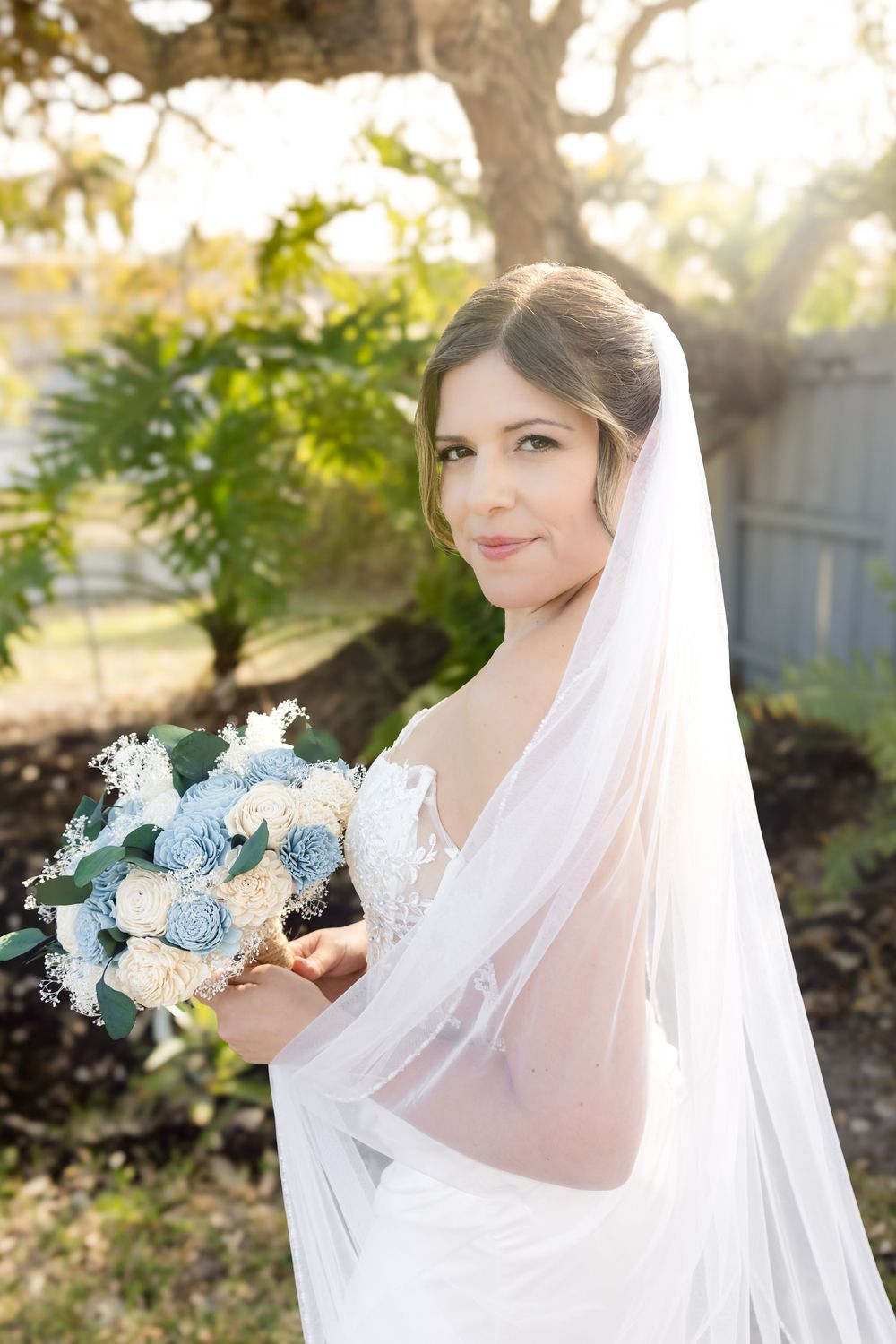 Bride in white gown and veil holds blue and ivory bouquet outdoors with sunlit greenery.