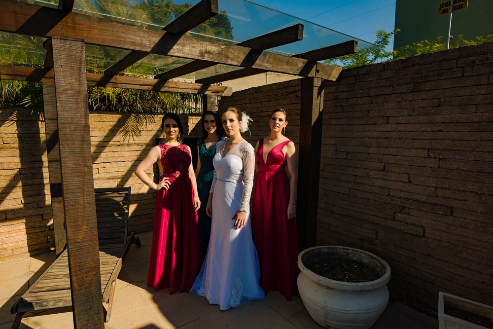 Three women in formal dresses standing under a pergola in bright sunlight.