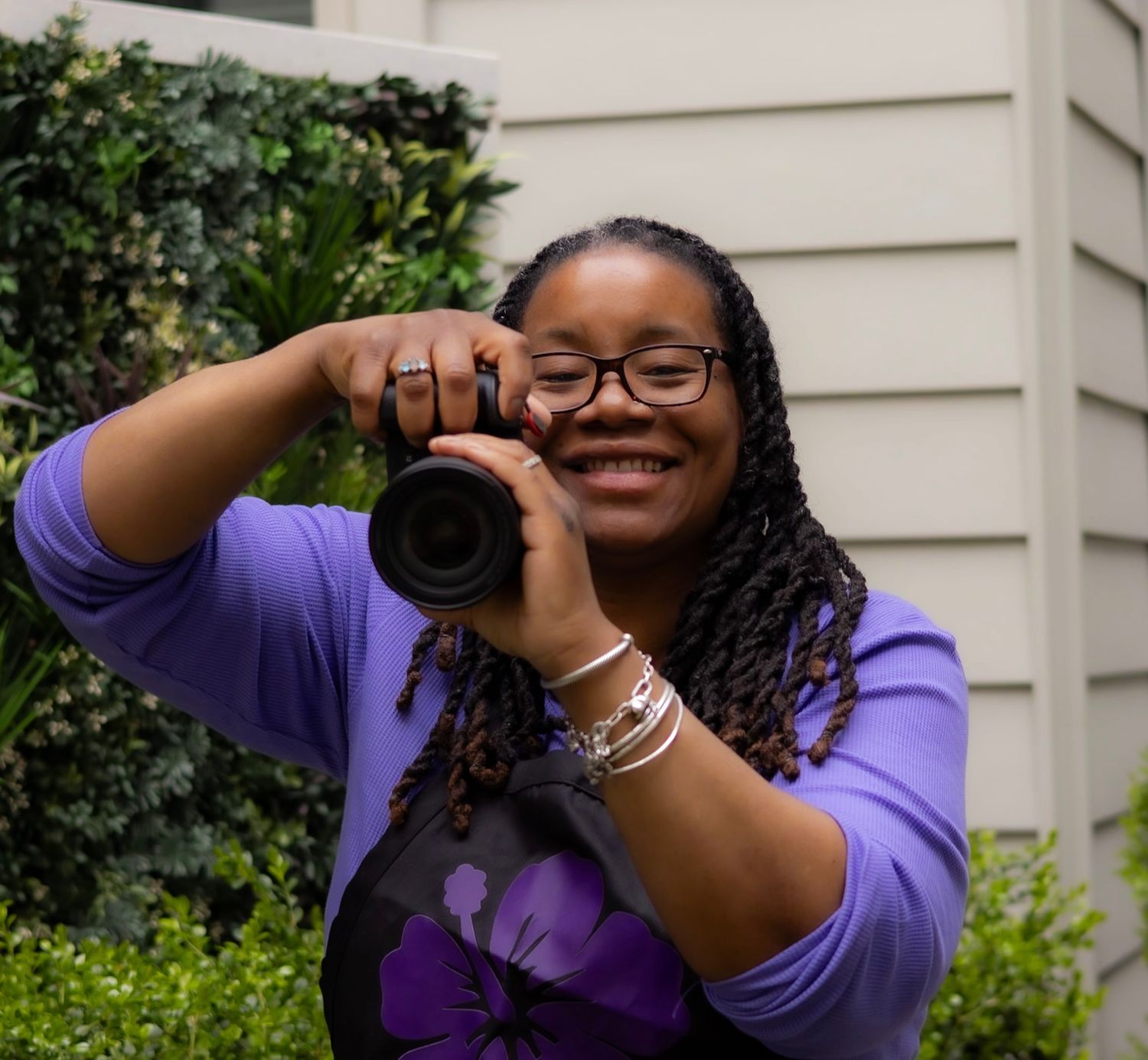 Smiling female photographer in purple hoodie holding DSLR camera outdoors among green foliage.