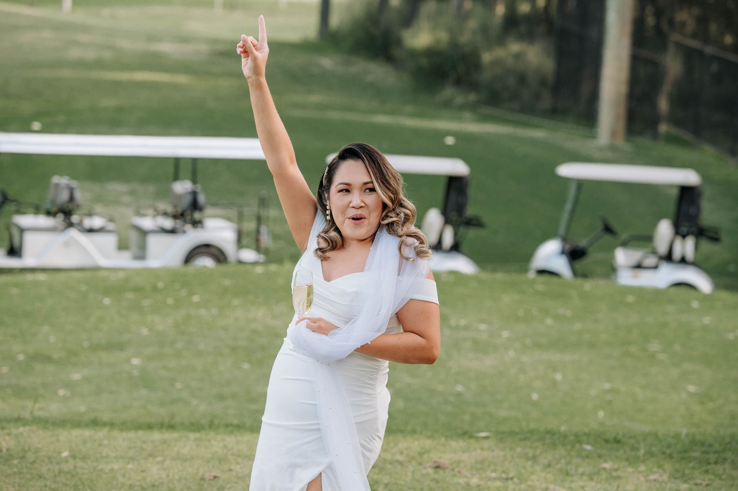Energetic bride in white dress dancing outdoors with golf carts visible in background.