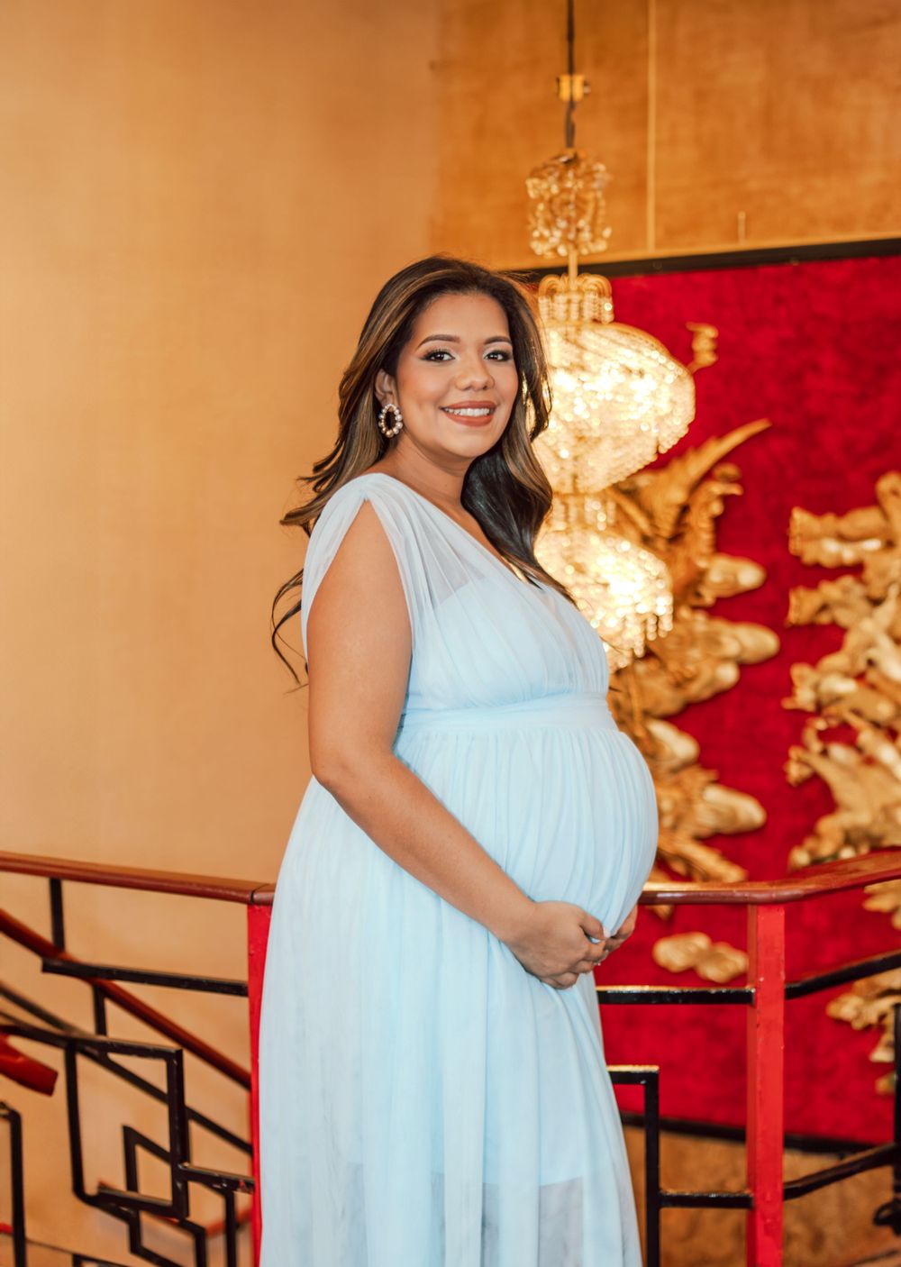 A woman in a light blue maternity dress poses in front of an ornate gold and red wall.