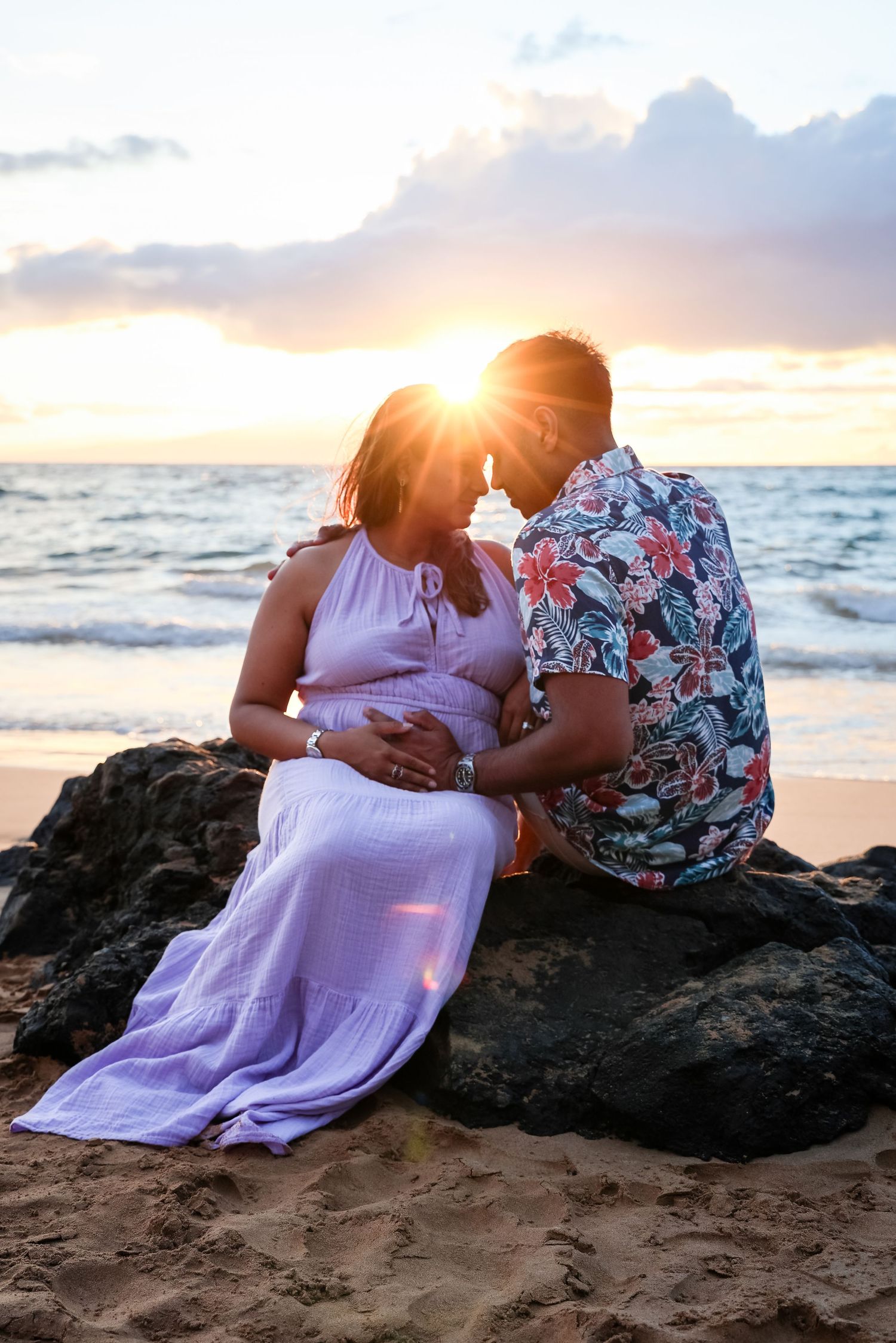 A couple sits together on black volcanic rocks at sunset on a beach in Hawaii during a romantic photoshoot.