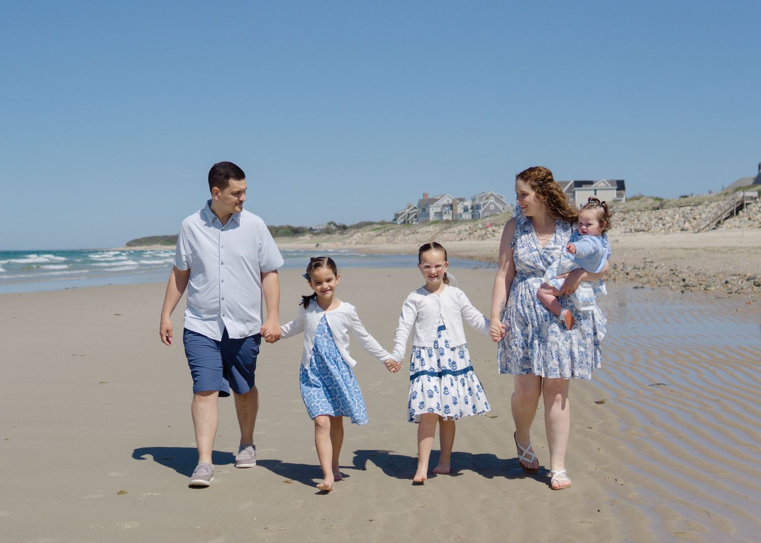 Family of four walking hand-in-hand along the water’s edge on a sunny Cape Cod beach day