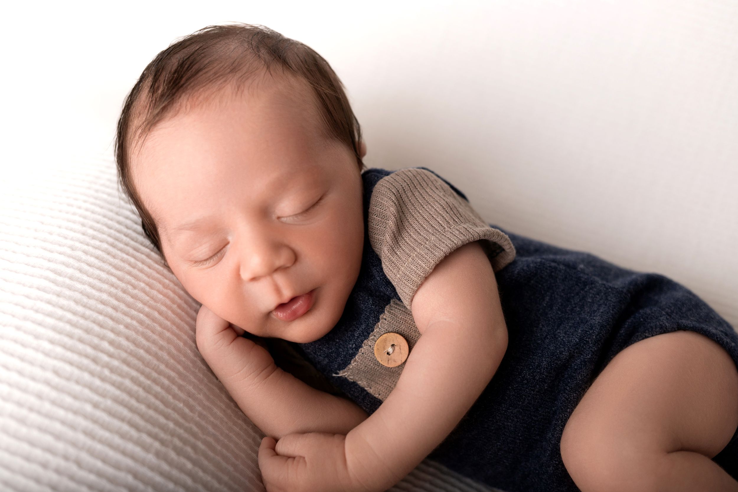 Peaceful sleeping newborn wearing navy and beige outfit on white pillow.