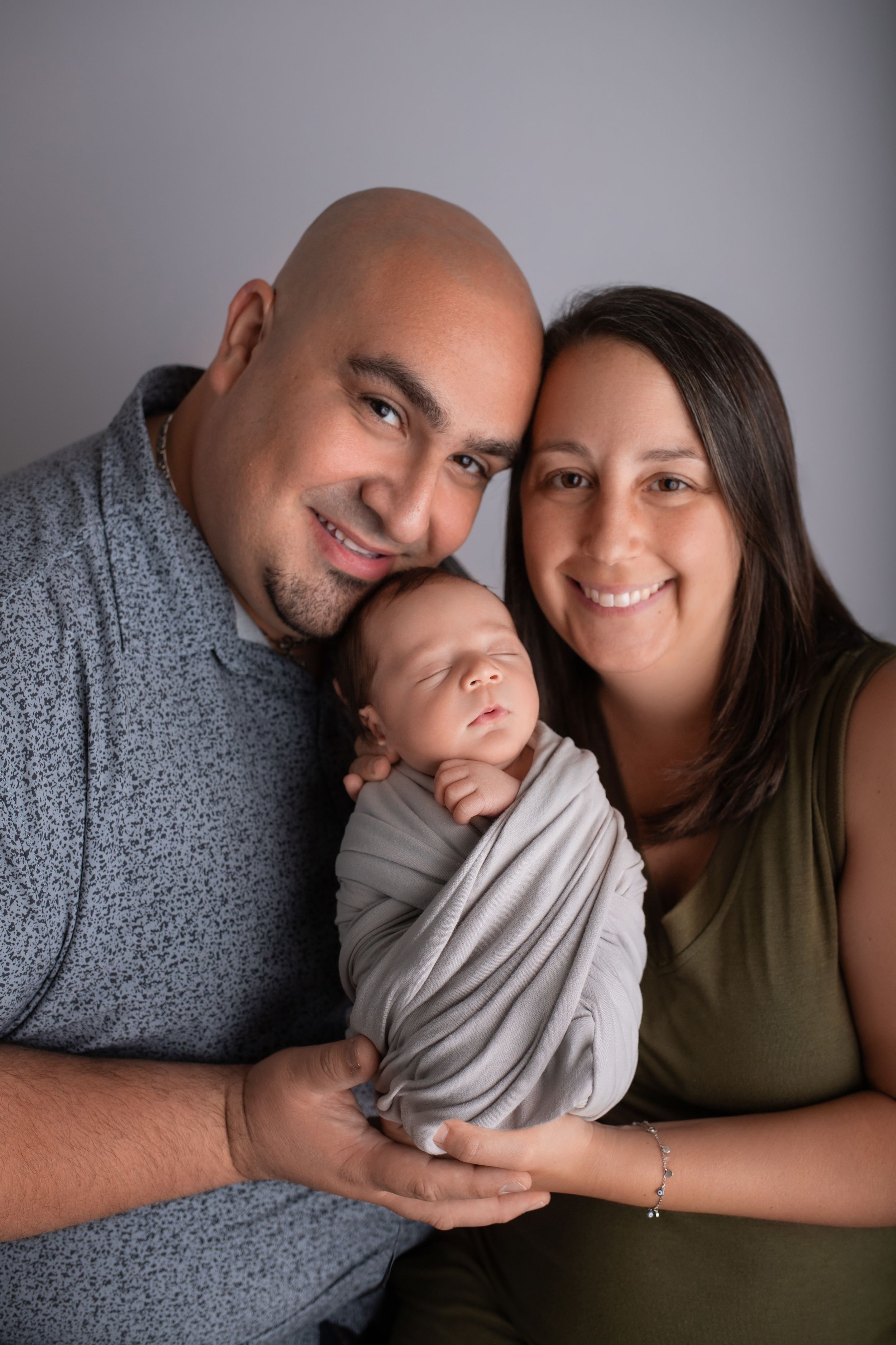 A loving family portrait with parents cradling their newborn baby wrapped in gray fabric.