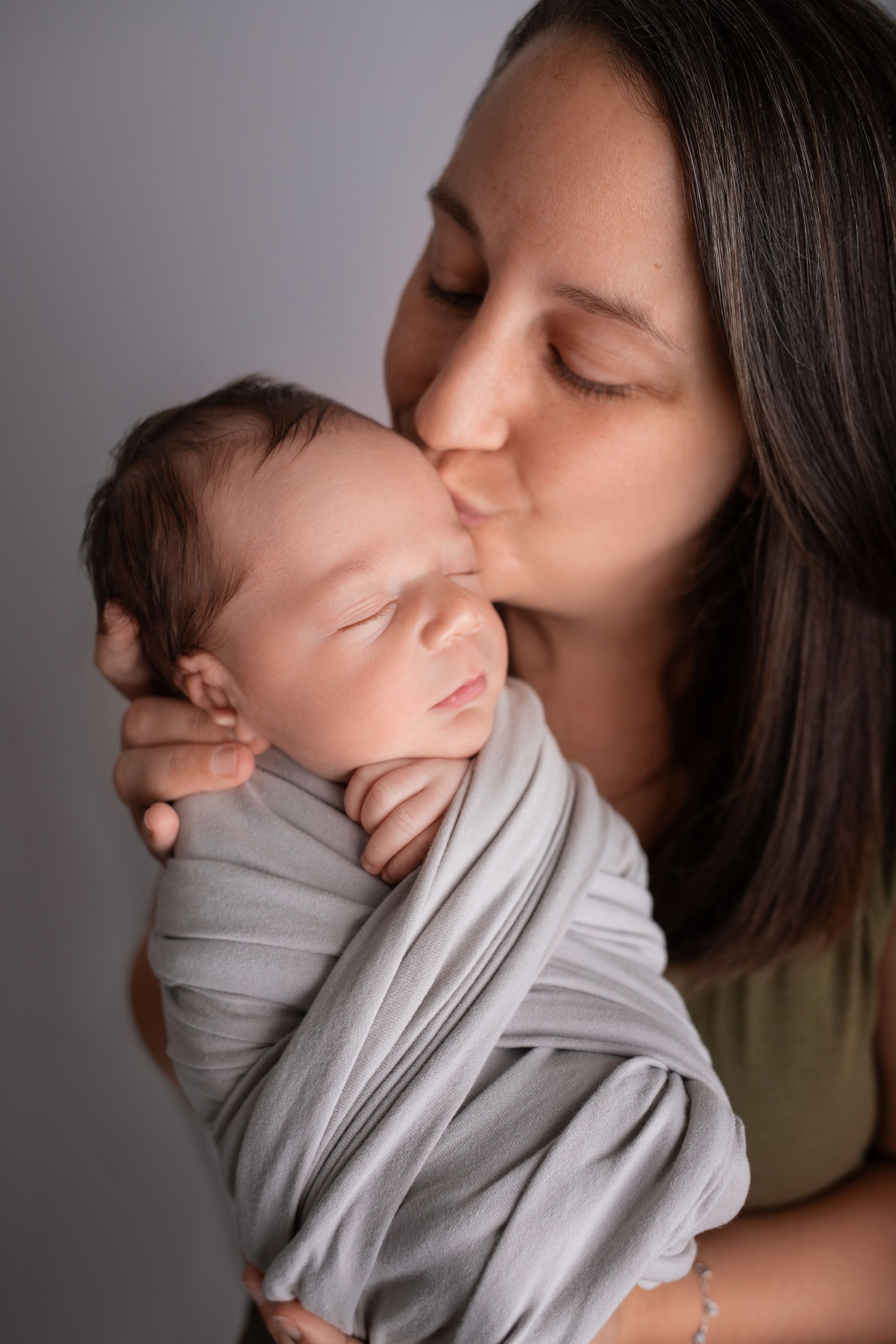 A gentle kiss shared between parent and sleeping newborn baby wrapped in soft gray fabric.