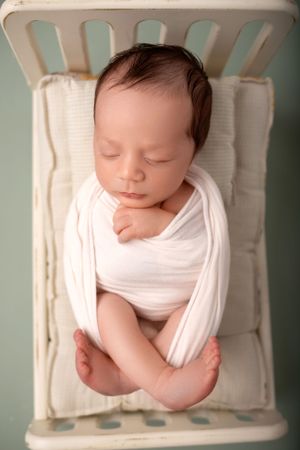 Peaceful sleeping newborn baby wrapped in white fabric lying on vintage-style chair.