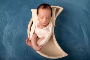 Sleeping newborn baby wrapped in white fabric lying in cream-colored prop on blue background.
