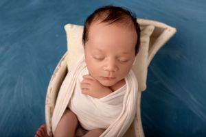 Peaceful sleeping newborn wrapped in white against vibrant blue backdrop.