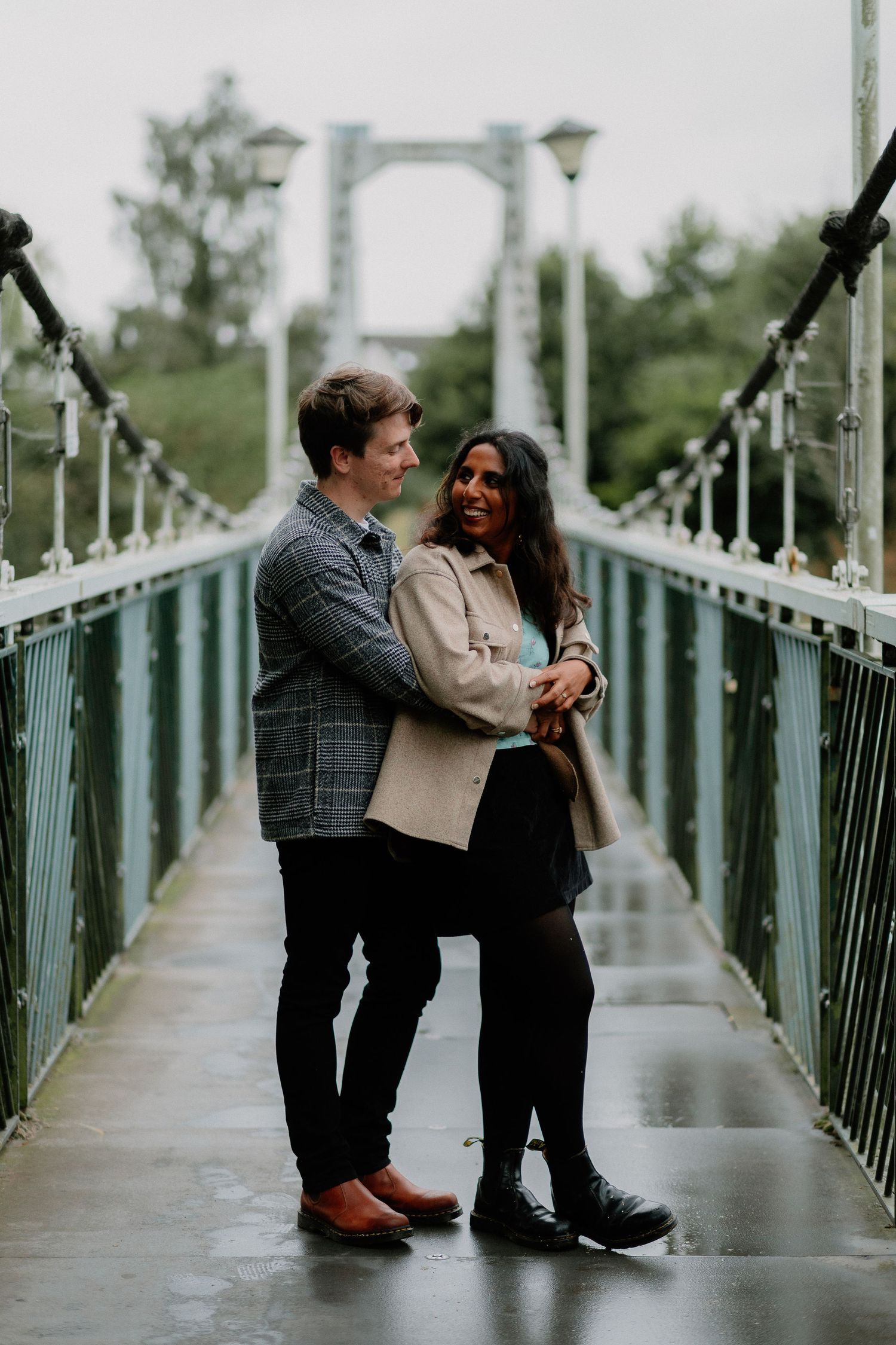 A couple embraces on a suspension bridge during an overcast day surrounded by greenery.