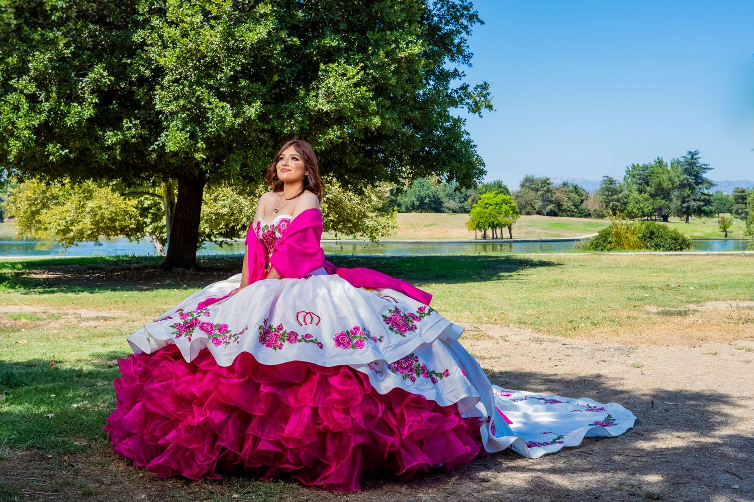 A vibrant magenta and white quinceañera dress with ruffled layers photographed in a sunny park setting.