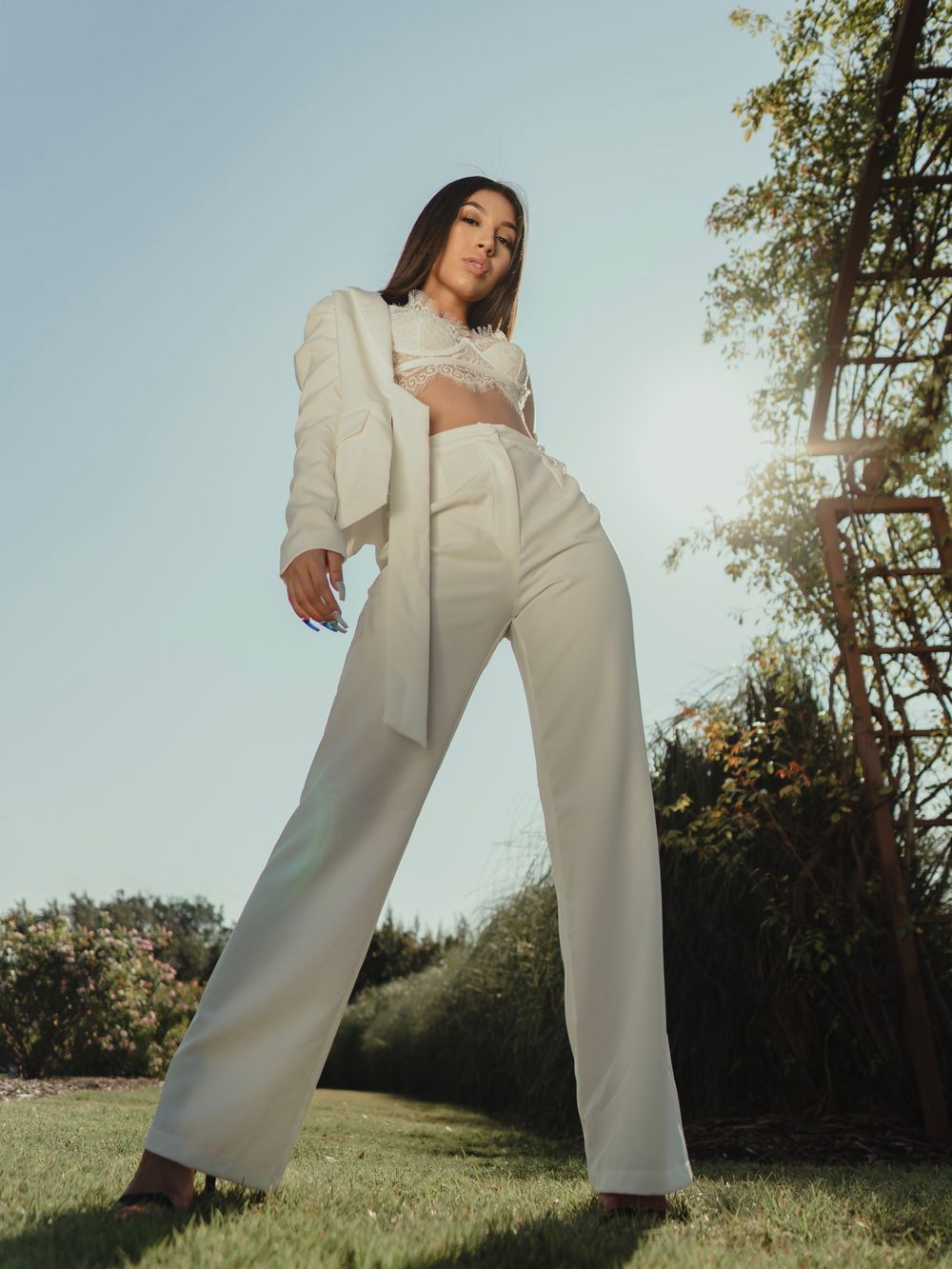 A model in an all-white pantsuit poses against a sunny outdoor backdrop.