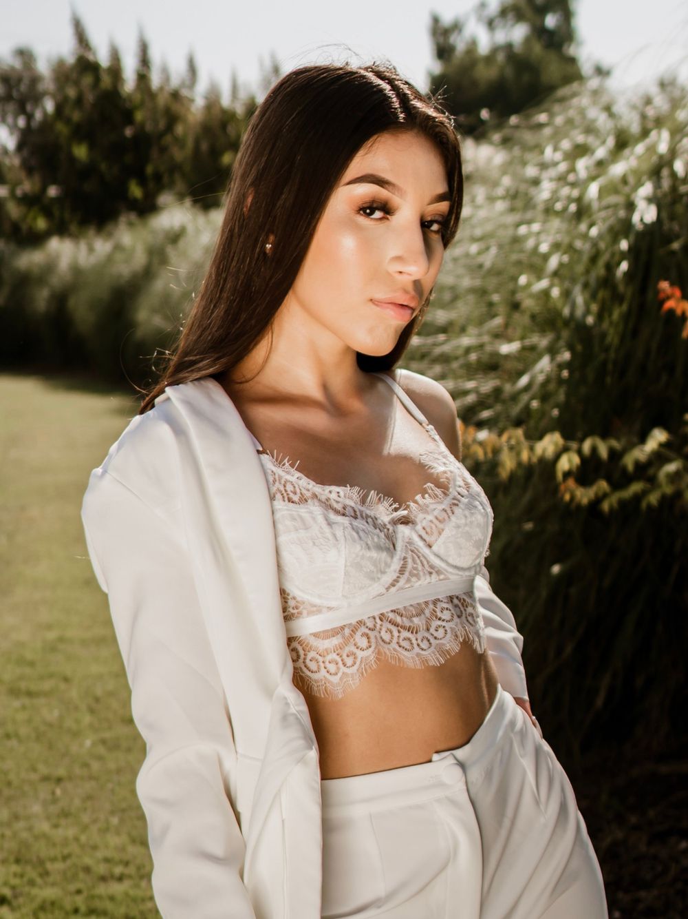 A model wears a white lace crop top and blazer in a sunny garden setting.