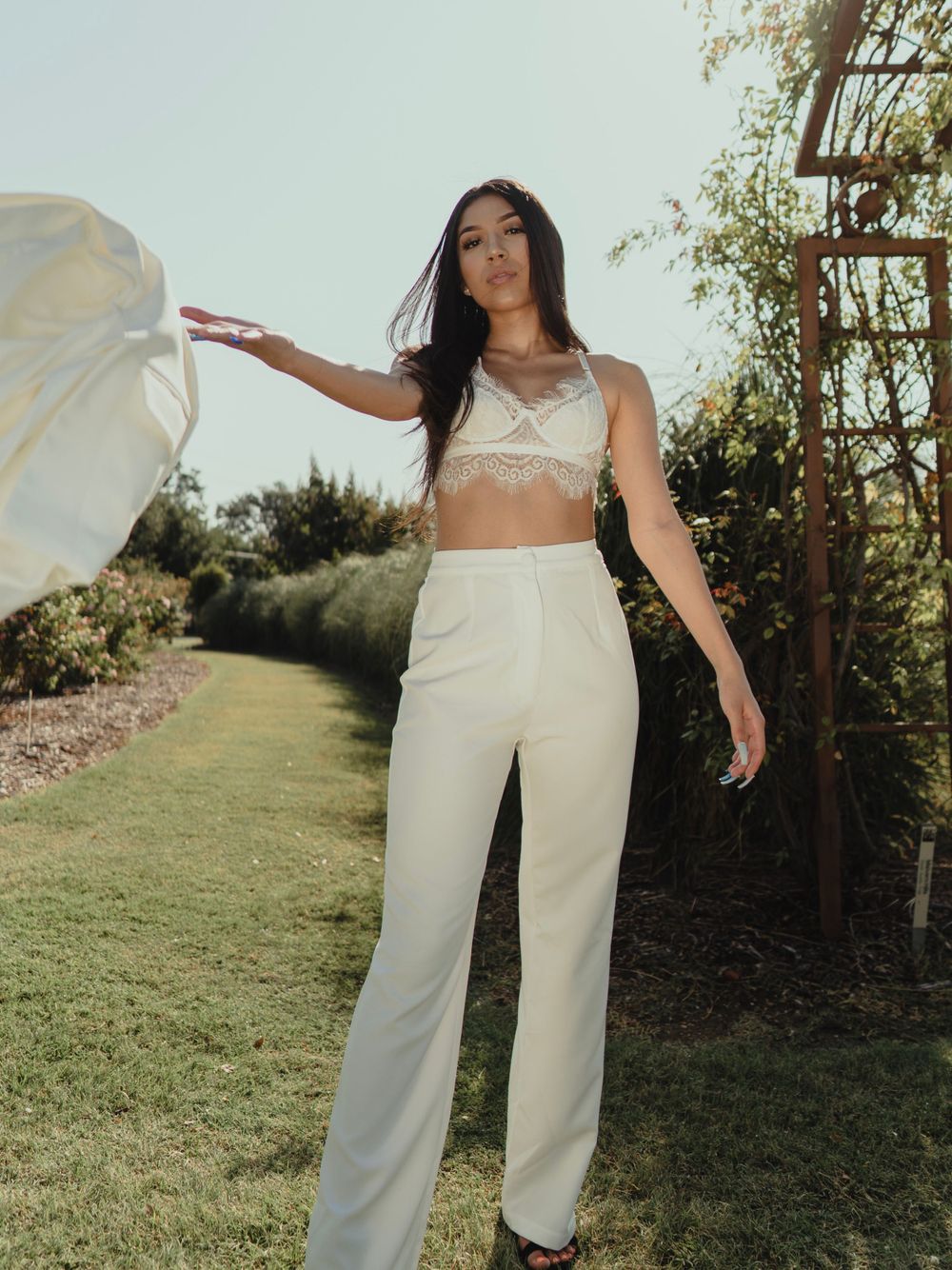 A model in white lace top and pants poses dramatically in a garden setting.