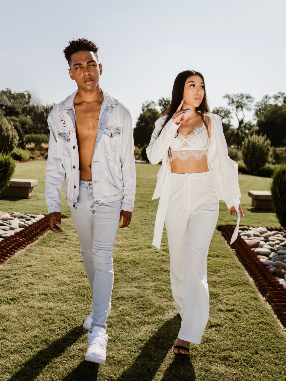 Fashion duo poses in coordinated light denim and white outfits on garden path.