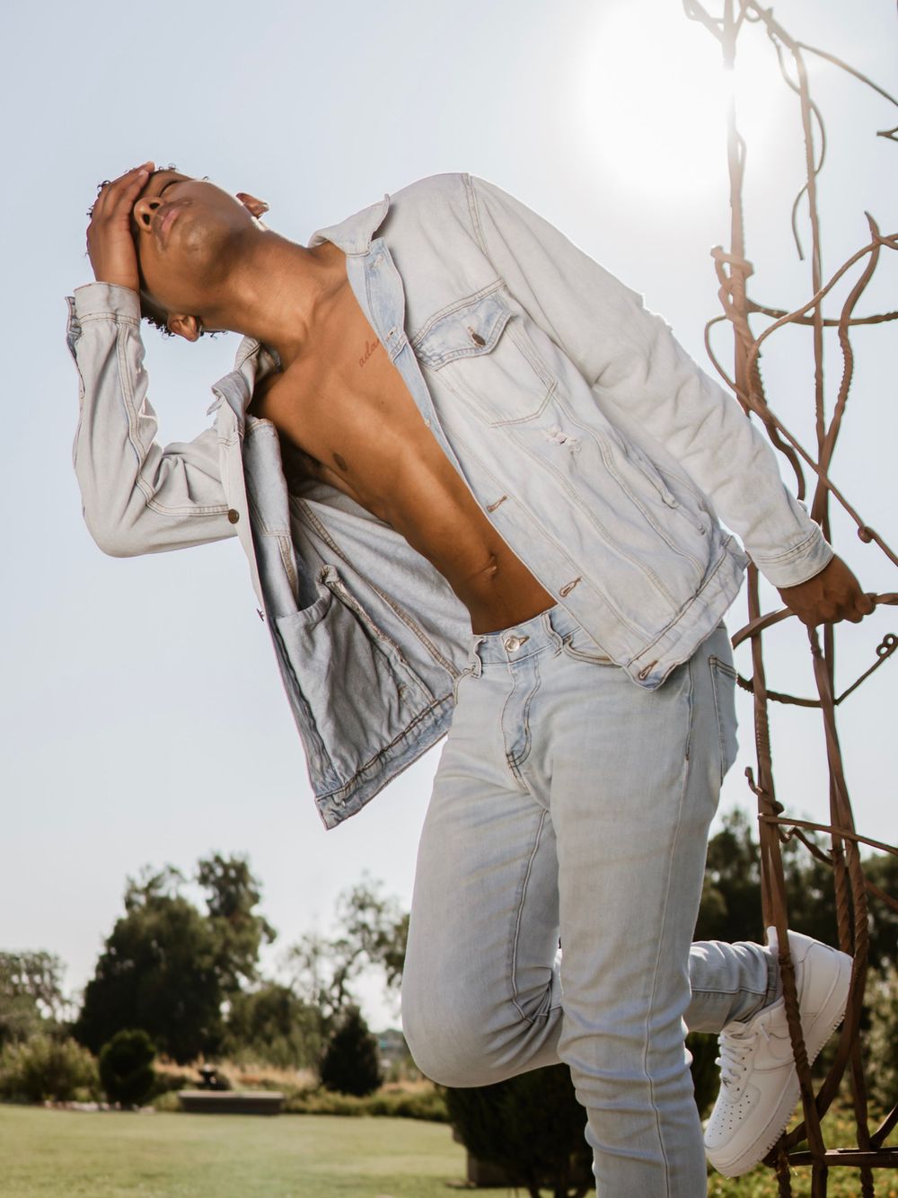 Model in light denim jacket and jeans poses dramatically in garden with sunlight.