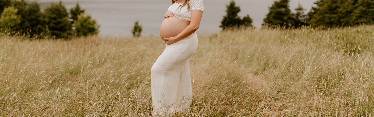 lady hugging her pregnant bare belly in a field with the ocean in the background