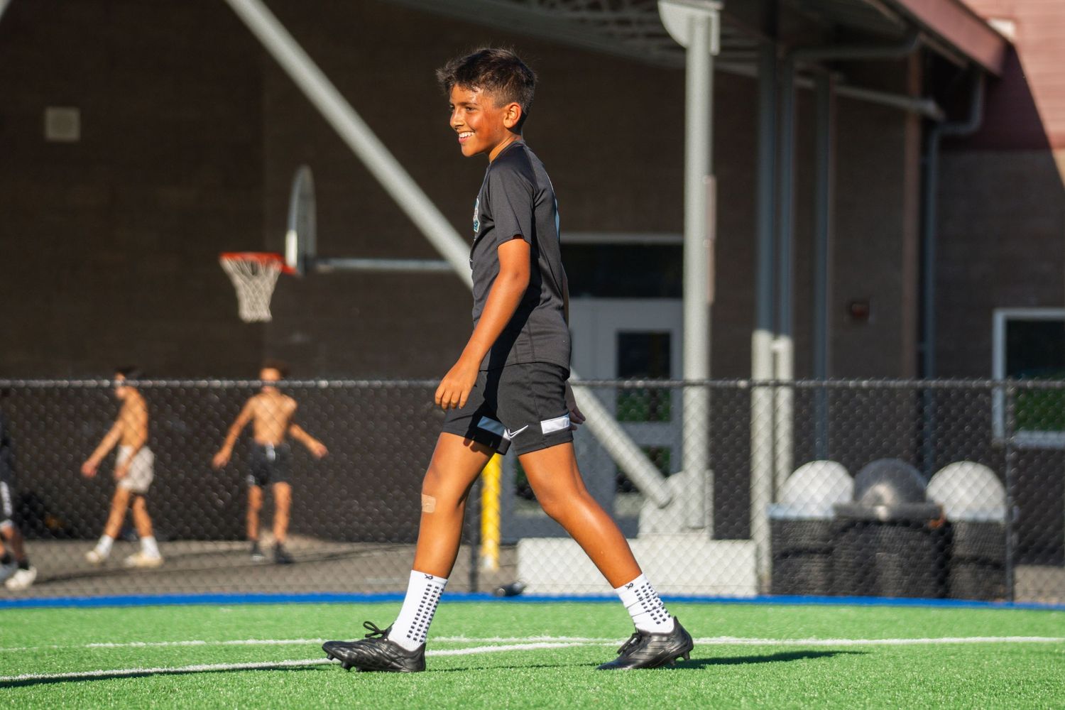 Young athlete in sports uniform walking across turf field with basketball court in background.