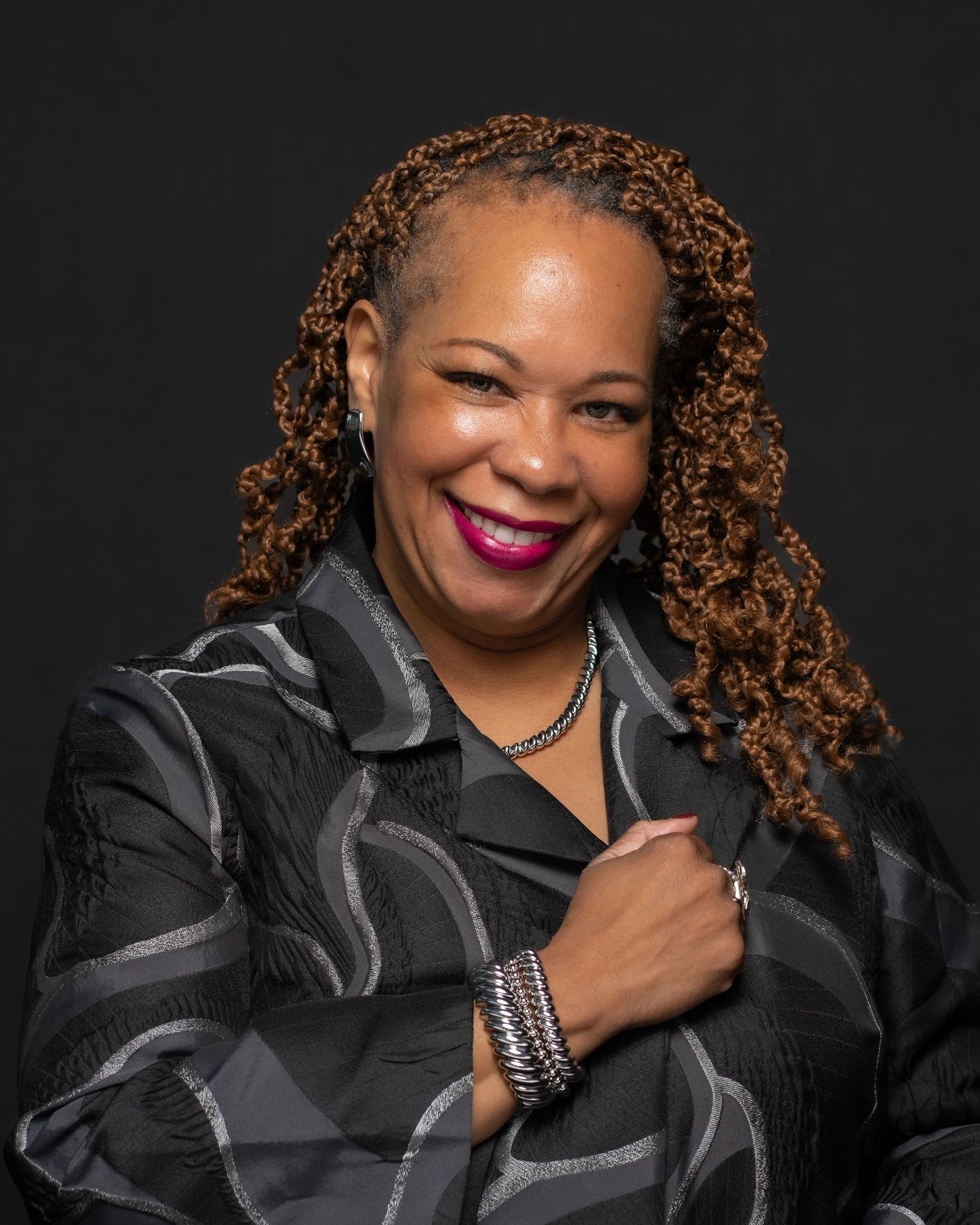 Professional headshot of smiling woman with curly locs, wearing a patterned black jacket and jewelry, posed confidently