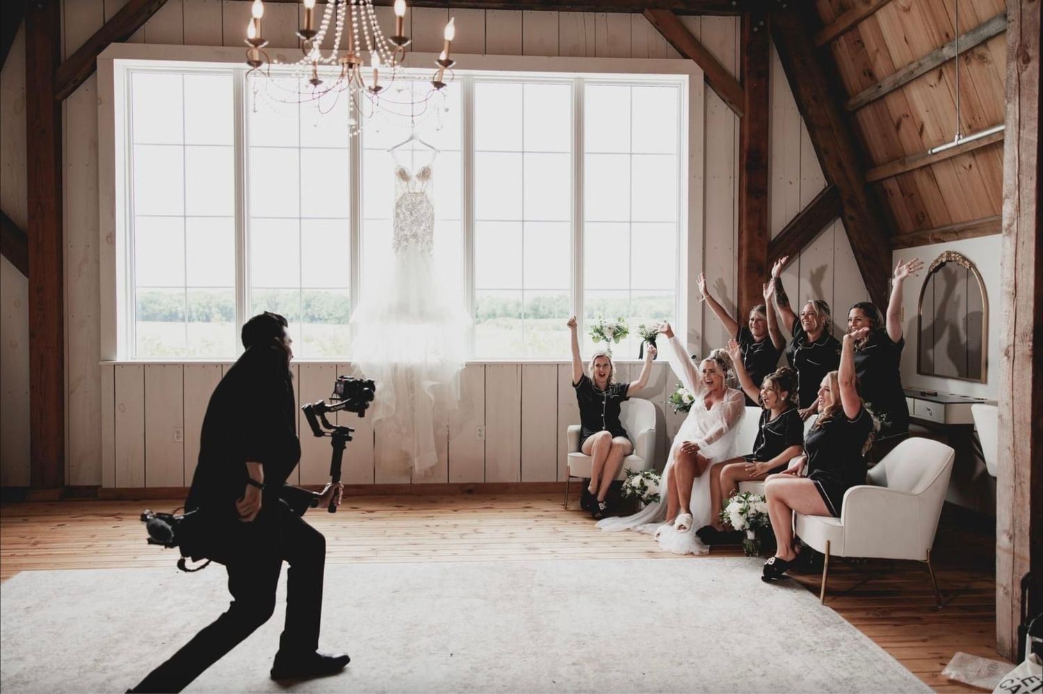 Wedding videographer Nate Templeton dressed in a black suit with gimbal and wedding dress hanging films excited bridesmaids on a white couch