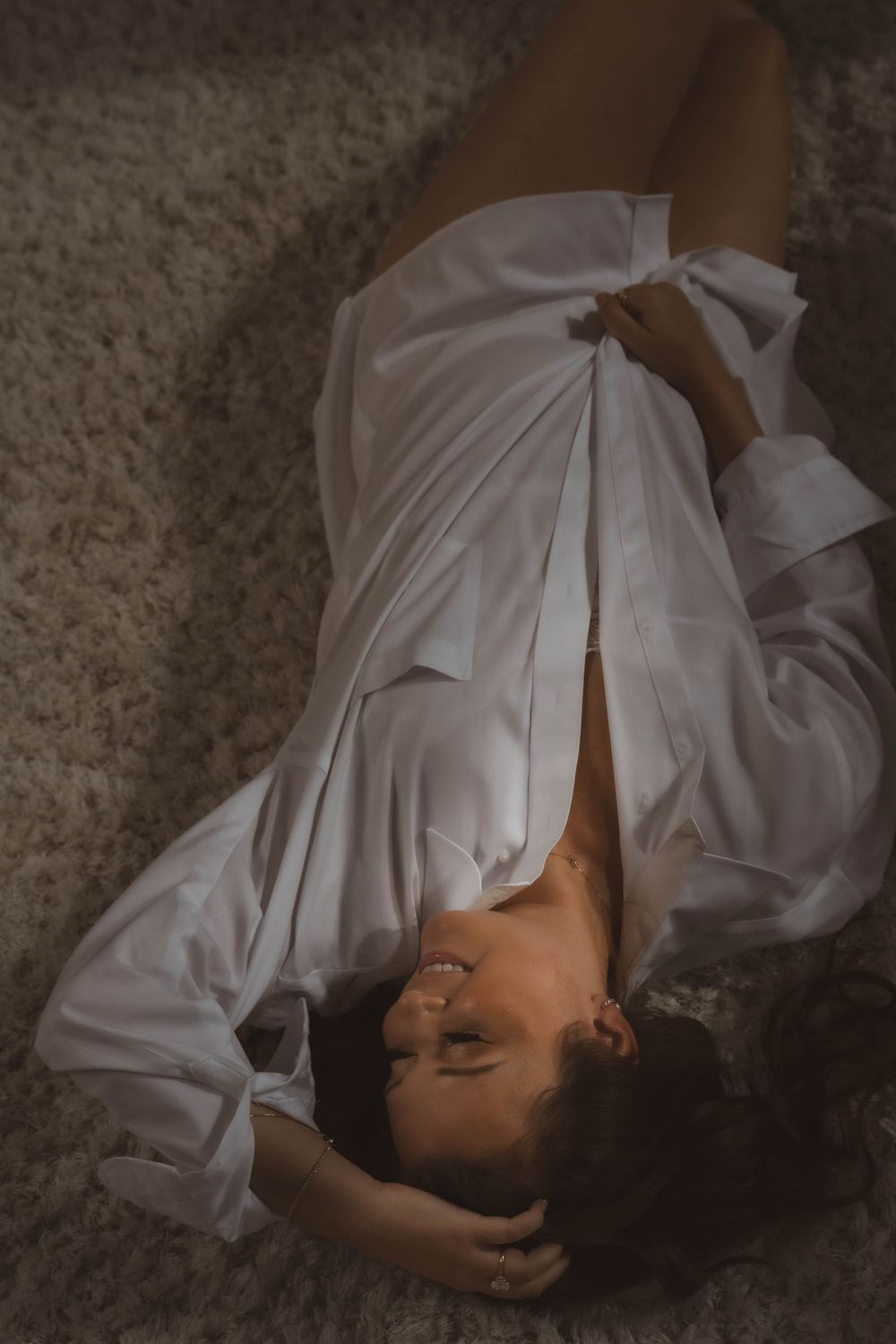 Smiling woman with dark hair lying on a soft shag carpet in an oversized white button-up shirt, in a moody boudoir-style portrait.