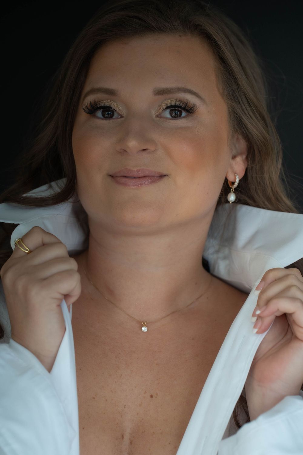 Smiling woman with long brown hair wearing a white collared shirt, pearl drop earrings, a delicate diamond pendant necklace and a gold ring.