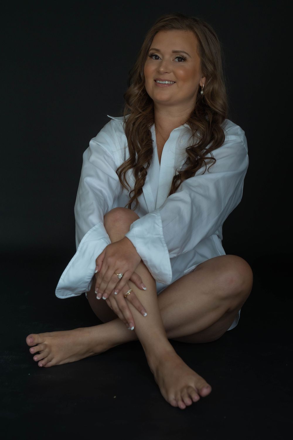 Smiling woman with long wavy auburn hair sits barefoot on the floor in a white button-up shirt against a dark studio background.