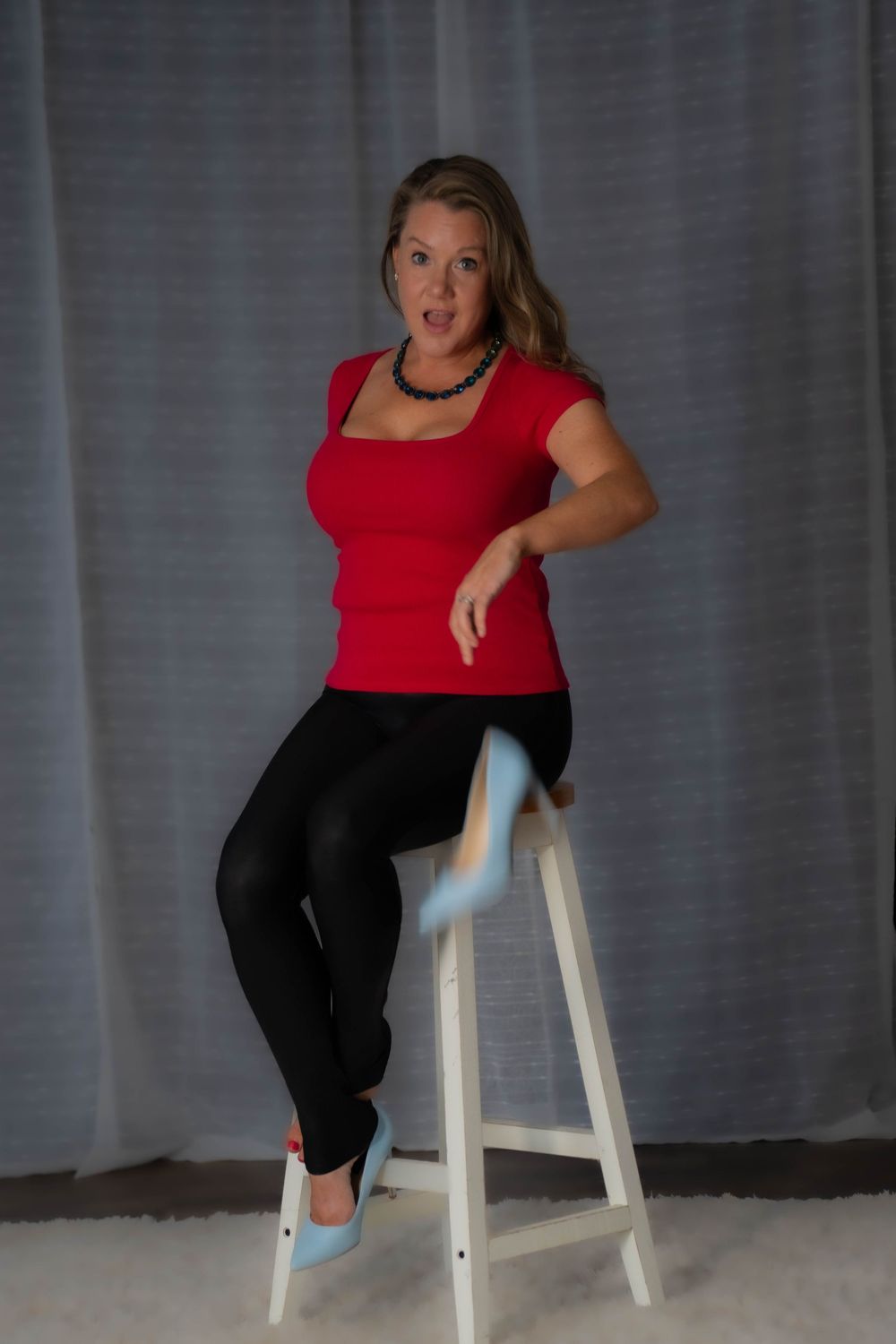 Person in red top and black pants sitting on white stool against grey backdrop.