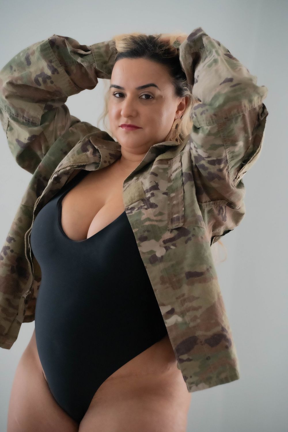 Person wearing black bodysuit and camouflage military jacket in studio setting.