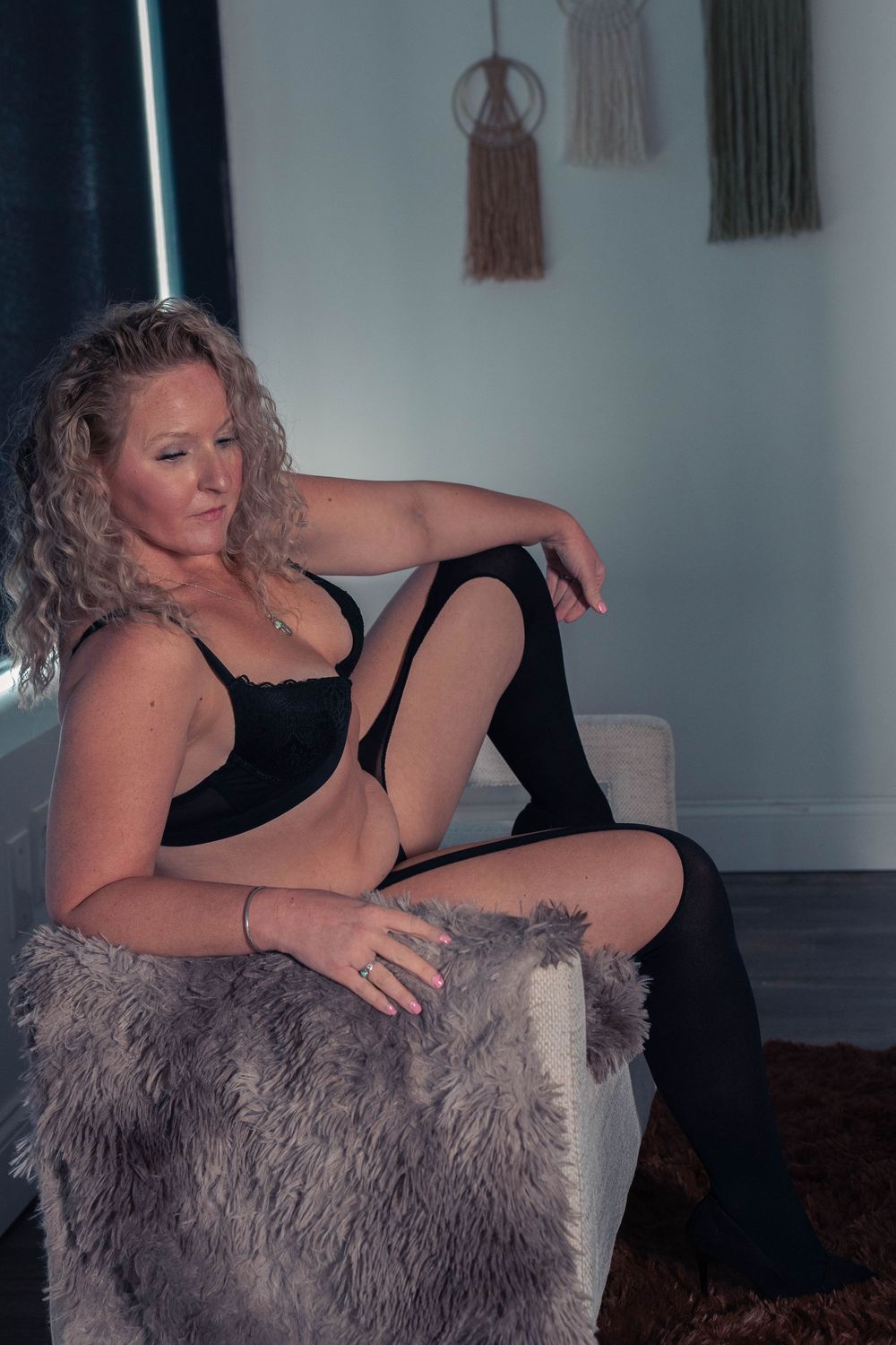 Person in black lingerie posing on furry chair with macrame wall decor.