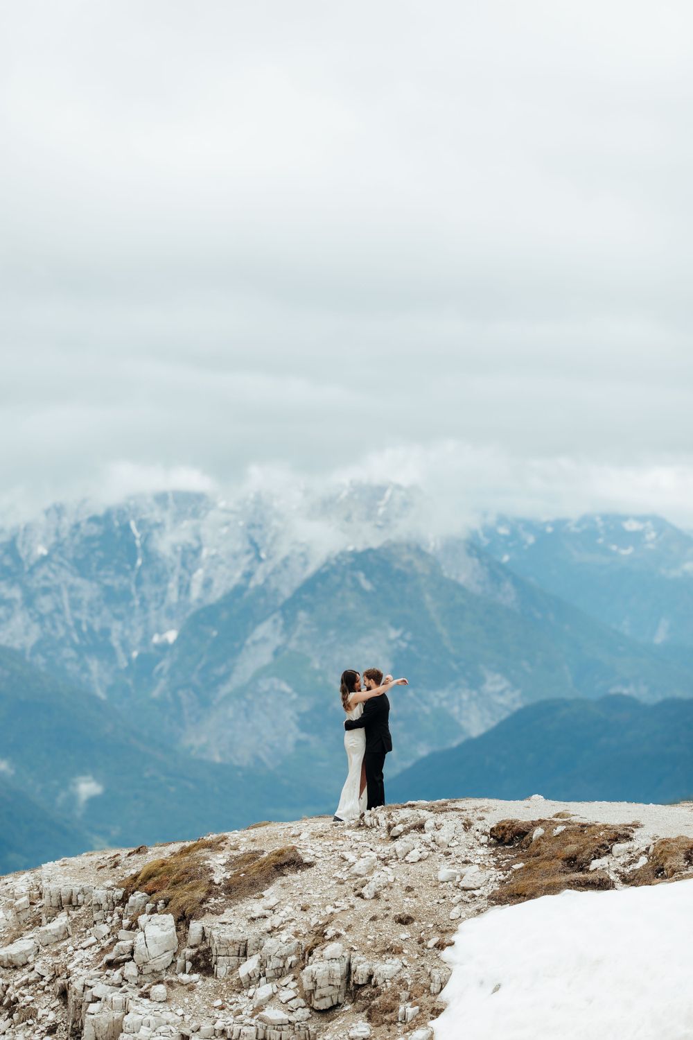 A couple embraces on a mountaintop with dramatic snow-covered peaks in the background.